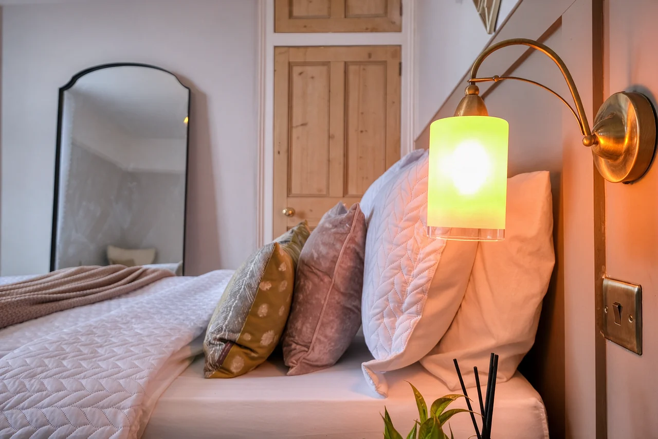 Close-up of a staged bedroom with layered cushions, a cosy throw and a warm bedside wall light